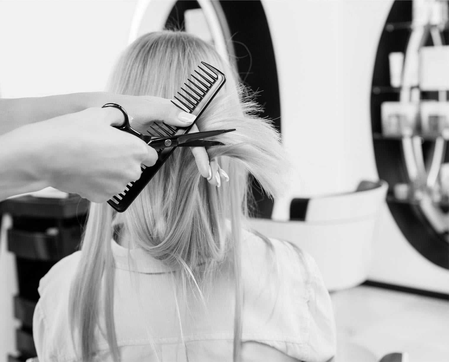 Hairdresser cutting client's long blonde hair with scissors and comb in salon.