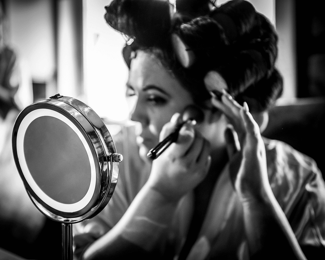 Woman applying makeup with curlers, reflected in illuminated mirror, black and white photo.
